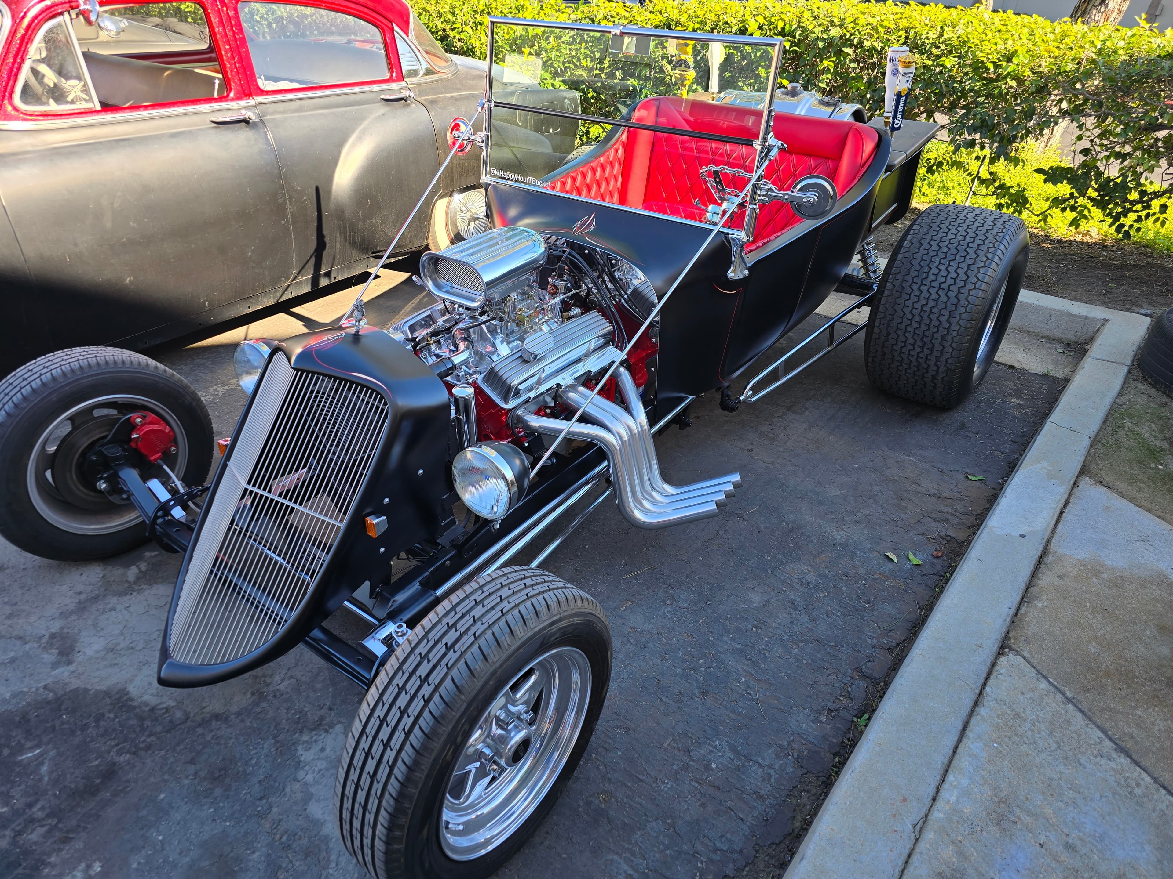 Matte black Ford T-bucket hot rod with red diamond-tufted interior and chrome blown V8 engine parked on a driveway